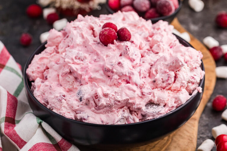 Pink whipped holiday dessert in a black bowl, garnished with sugared cranberries, placed on a wooden board with scattered mini marshmallows, frosted berries, and a red and green plaid napkin.