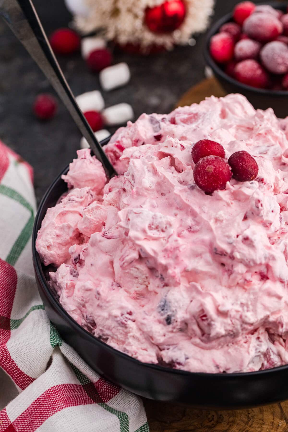 Close-up of a pink whipped holiday dessert in a black bowl, garnished with sugared cranberries and a serving spoon, set beside a red and green plaid napkin with festive decor and mini marshmallows in the background.