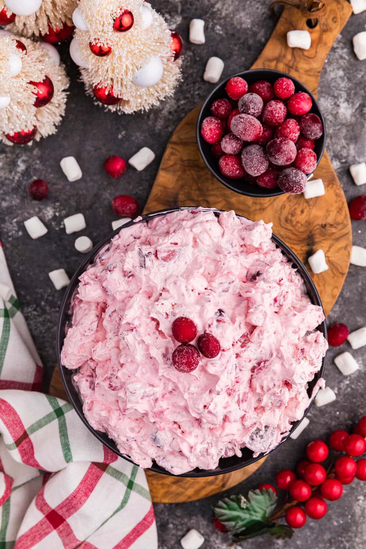 Pink whipped dessert topped with sugared cranberries in a black bowl, surrounded by mini marshmallows, frosted berries, holiday decorations, and a red and green plaid napkin on a rustic dark surface.