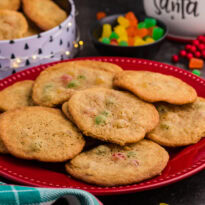 Holiday cookies with colorful gumdrops arranged on a red plate, surrounded by Christmas-themed decor, a candy bowl, and a festive cookie tin in the background.