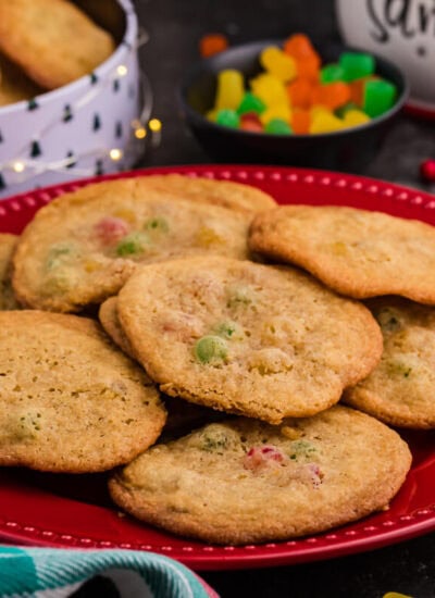 Holiday cookies with colorful gumdrops arranged on a red plate, surrounded by Christmas-themed decor, a candy bowl, and a festive cookie tin in the background.