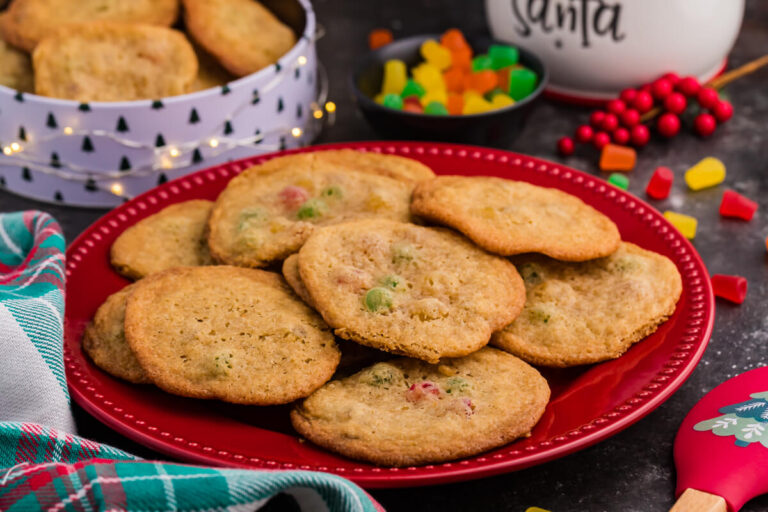 Holiday cookies with colorful gumdrops arranged on a red plate, surrounded by Christmas-themed decor, a candy bowl, and a festive cookie tin in the background.