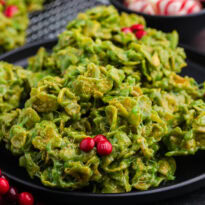 Close-up of green cornflake wreath cookies on a black plate, with red M&M candies as holly berries. Background details include peppermint swirl chocolates in a bowl and festive red berry decorations.