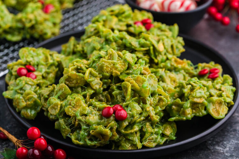 Close-up of green cornflake wreath cookies on a black plate, with red M&M candies as holly berries. Background details include peppermint swirl chocolates in a bowl and festive red berry decorations.