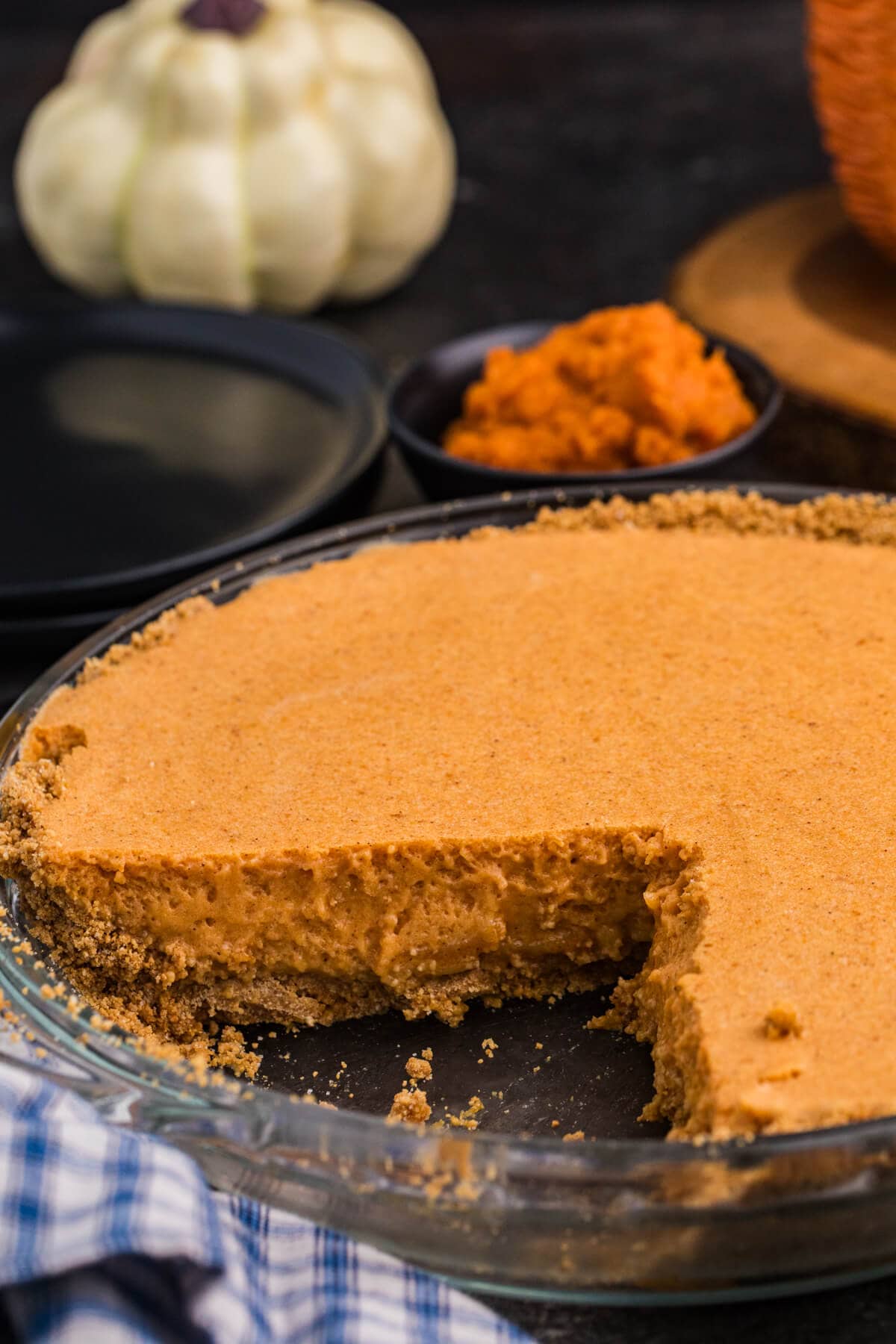 A partially sliced creamy pumpkin pie sits in a glass pie dish with a graham cracker crust, with pumpkins and a bowl of pumpkin purée in the background.