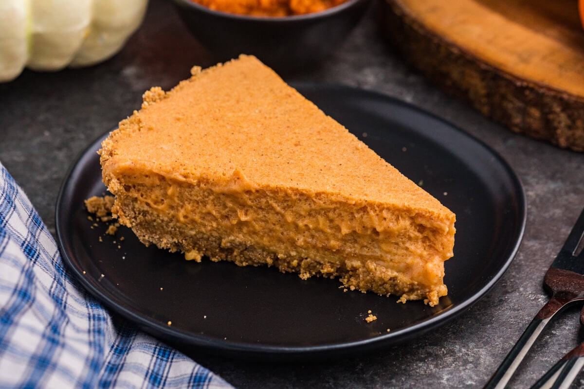 A plain slice of chilled pumpkin pie with a graham cracker crust sits on a black plate with pumpkins and a bowl of pumpkin purée in the background.