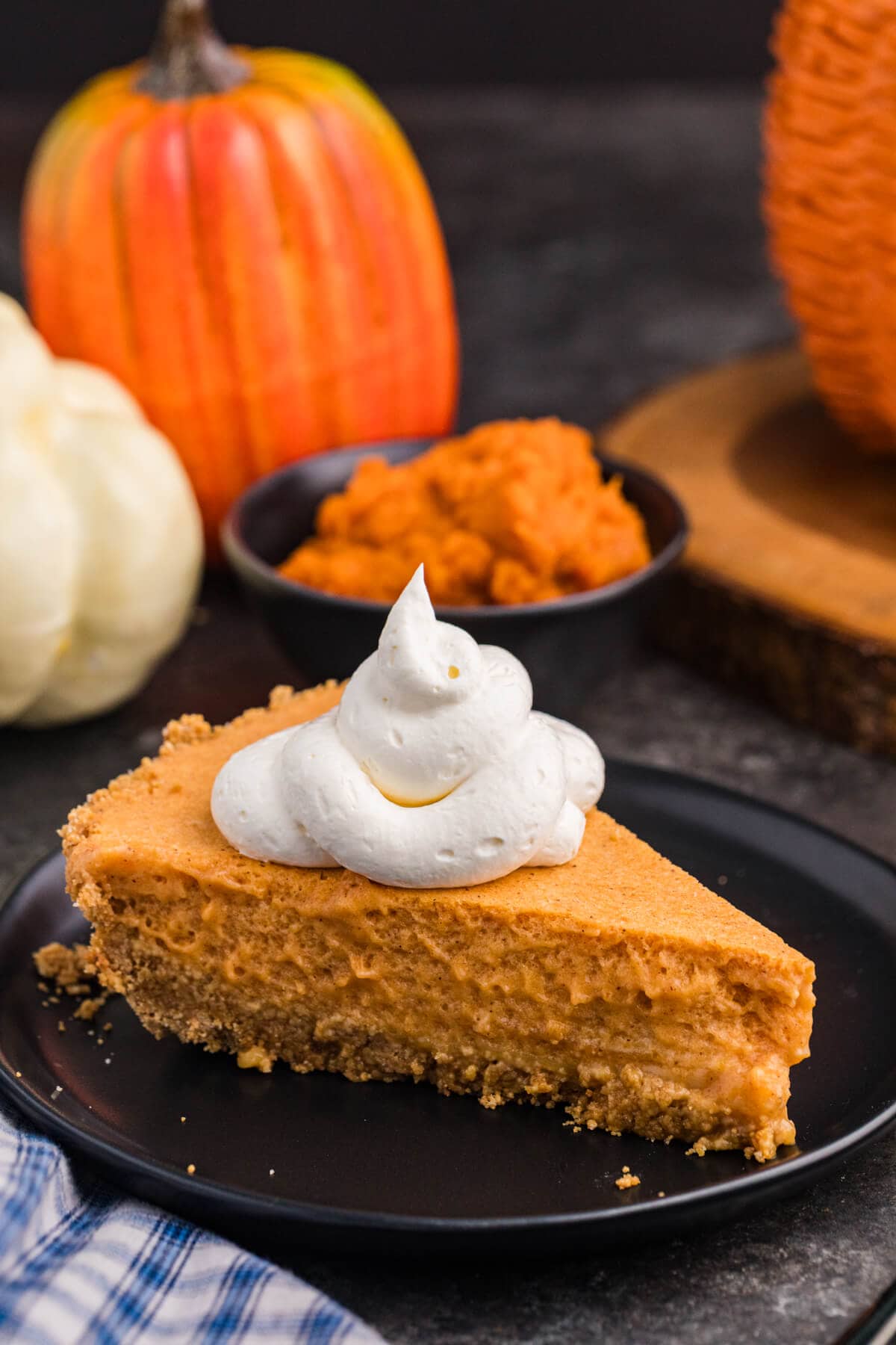 A slice of pumpkin chiffon pie topped with whipped cream sits on a black plate with pumpkins and a bowl of pumpkin purée in the background.