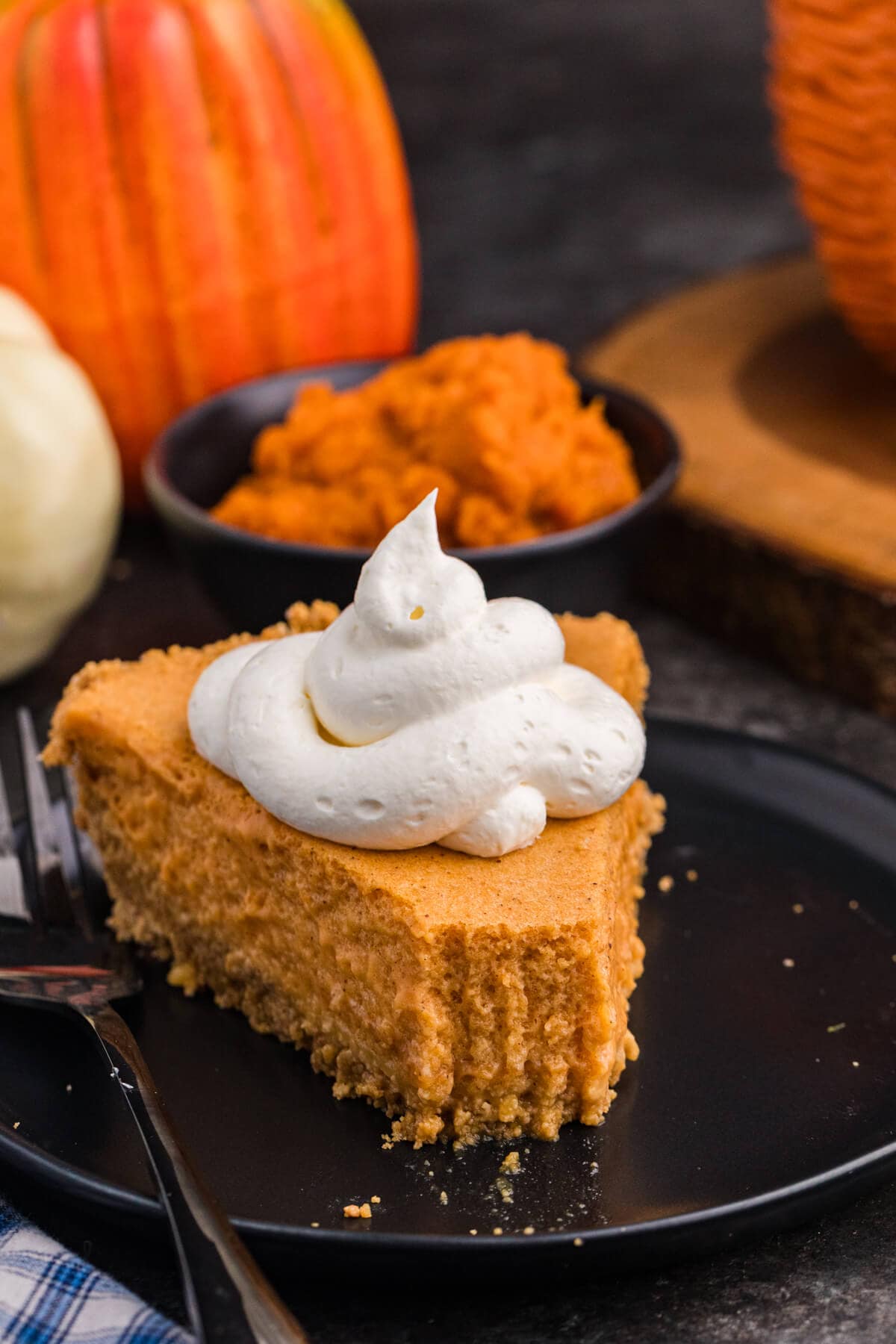 A slice of light, creamy pumpkin pie topped with a swirl of whipped cream sits on a black plate, with pumpkins and a bowl of pumpkin purée blurred in the background.