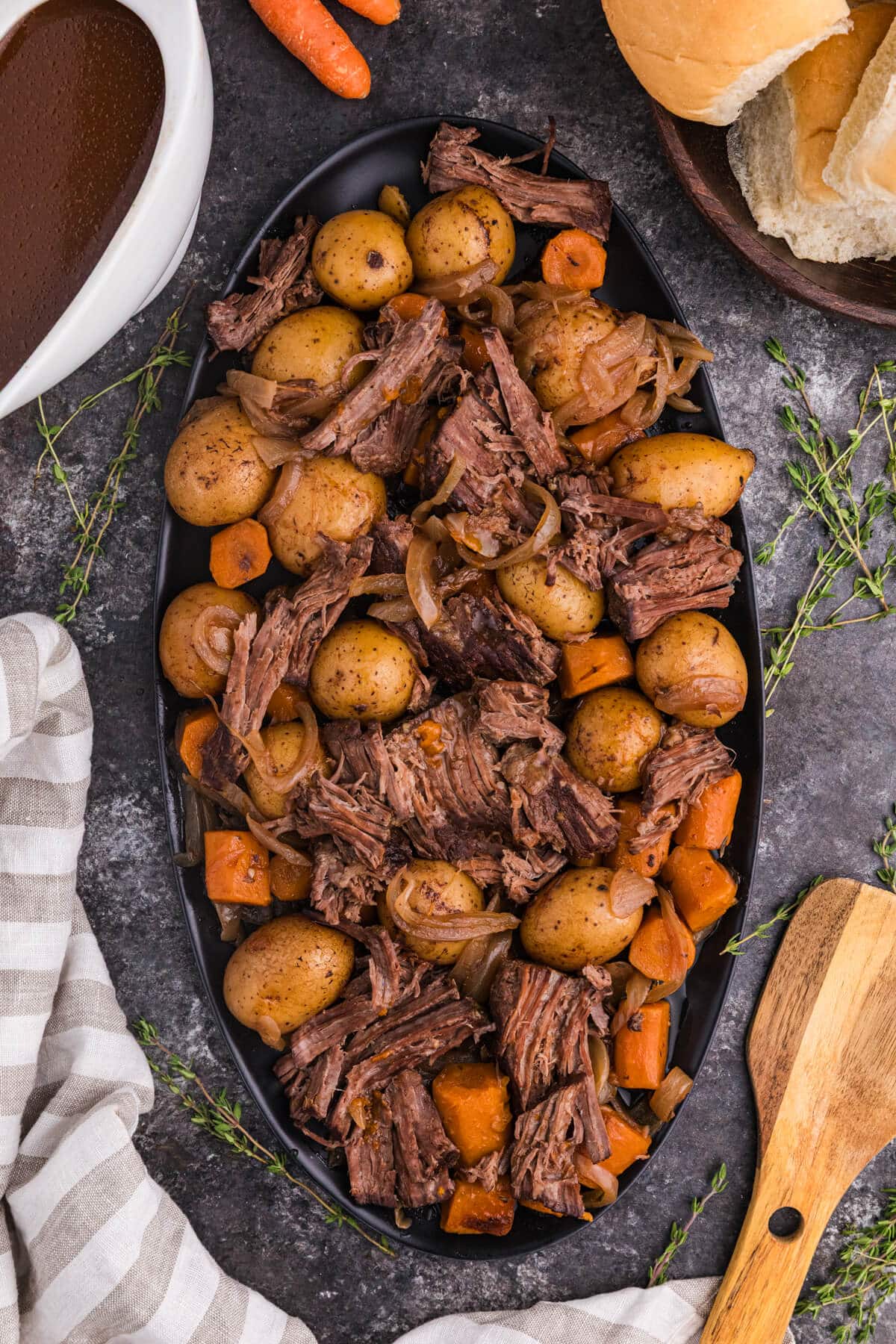 A serving platter filled with tender shredded slow cooker pot roast, baby potatoes, carrots, and onions, surrounded by a bowl of rich brown gravy, fresh thyme sprigs, dinner rolls, and a wooden spoon on a dark surface.