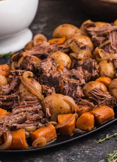 Shredded beef, baby potatoes, carrots, and onions arranged on a black serving platter with a bowl of gravy, fresh thyme, and a striped napkin beside it.