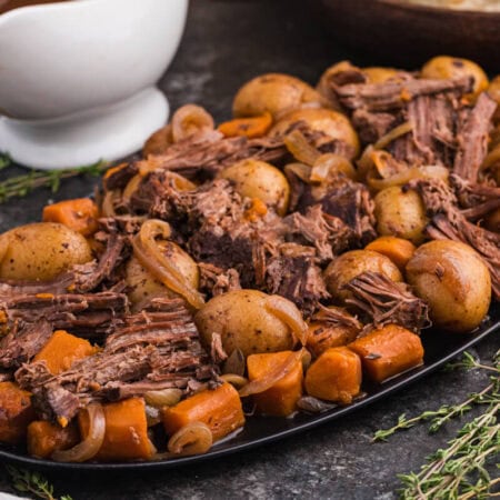 Shredded beef, baby potatoes, carrots, and onions arranged on a black serving platter with a bowl of gravy, fresh thyme, and a striped napkin beside it.