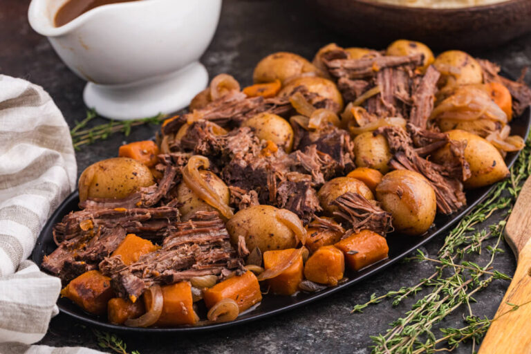 Shredded beef, baby potatoes, carrots, and onions arranged on a black serving platter with a bowl of gravy, fresh thyme, and a striped napkin beside it.