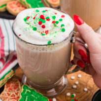 Close-up of a hand with red nail polish holding a festive glass mug filled with latte topped with whipped cream and red, green, and white holiday sprinkles, surrounded by decorated Christmas cookies and colorful sugar sprinkles on a wooden board.