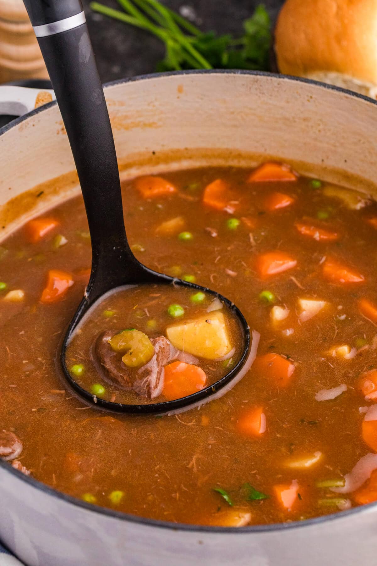 Close-up of hearty beef stew in a Dutch oven with a ladle, featuring beef chunks, potatoes, carrots, peas, and celery in a rich brown broth.