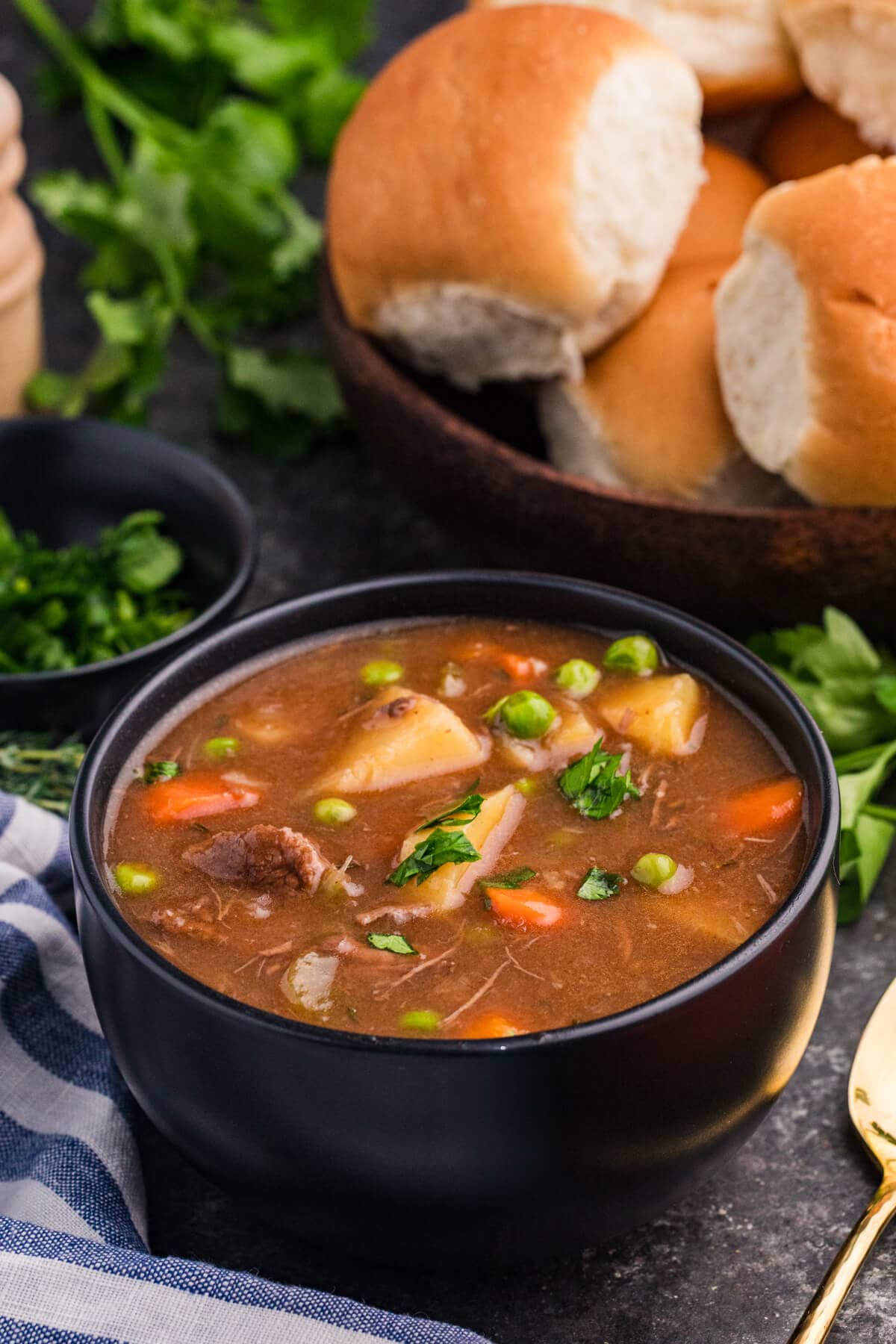 Bowl of meat and vegetable soup with potatoes, carrots, peas, and herbs, served with dinner rolls and fresh parsley.