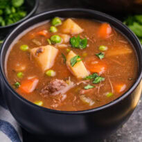 Side view of a bowl of stew with beef, carrots, potatoes, peas, and parsley, next to a towel and herbs.