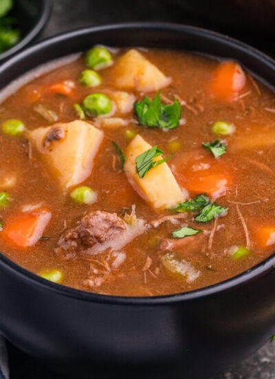 Side view of a bowl of stew with beef, carrots, potatoes, peas, and parsley, next to a towel and herbs.