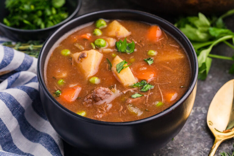 Side view of a bowl of stew with beef, carrots, potatoes, peas, and parsley, next to a towel and herbs.