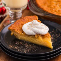 Slice of golden custard pie topped with whipped cream and nutmeg, served on a black plate with festive holiday decor and a glass of eggnog in the background.