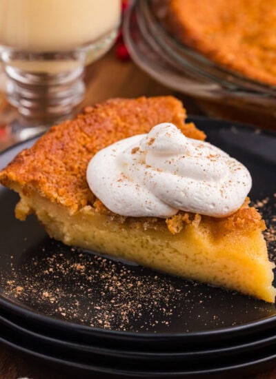 Slice of golden custard pie topped with whipped cream and nutmeg, served on a black plate with festive holiday decor and a glass of eggnog in the background.