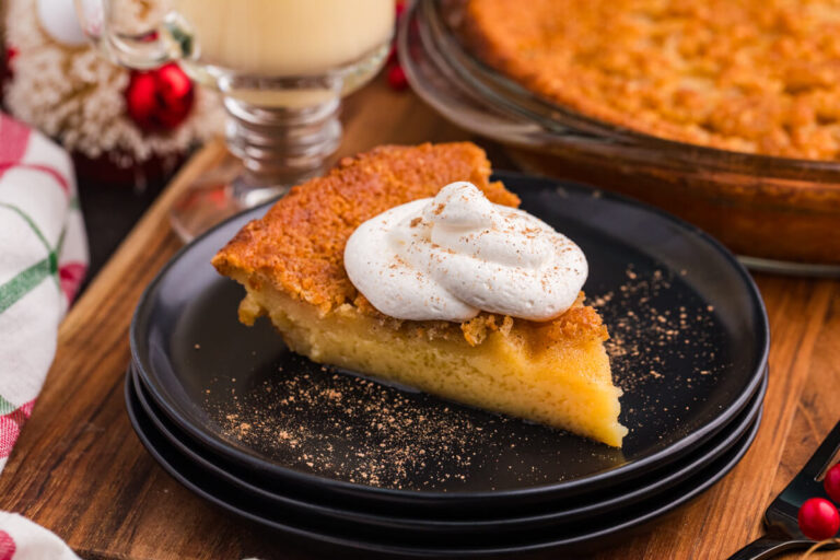 Slice of golden custard pie topped with whipped cream and nutmeg, served on a black plate with festive holiday decor and a glass of eggnog in the background.