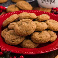 Plate of golden, sugar-coated holiday cookies stacked on a red dish, surrounded by festive decor including red berries and a “Cookies for Santa” jar in the background.
