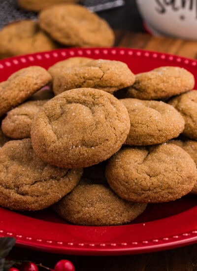 Plate of golden, sugar-coated holiday cookies stacked on a red dish, surrounded by festive decor including red berries and a “Cookies for Santa” jar in the background.