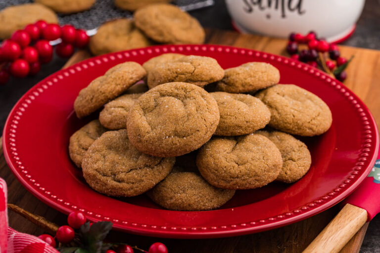 Plate of golden, sugar-coated holiday cookies stacked on a red dish, surrounded by festive decor including red berries and a “Cookies for Santa” jar in the background.