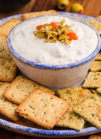 Close-up of a ceramic bowl filled with creamy white dip topped with chopped green and red pimento-stuffed olives, surrounded by seasoned multigrain and herb-flavored square crackers on a rustic serving plate.