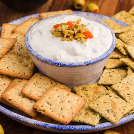 Close-up of a ceramic bowl filled with creamy white dip topped with chopped green and red pimento-stuffed olives, surrounded by seasoned multigrain and herb-flavored square crackers on a rustic serving plate.