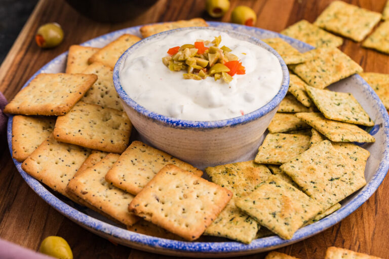 Close-up of a ceramic bowl filled with creamy white dip topped with chopped green and red pimento-stuffed olives, surrounded by seasoned multigrain and herb-flavored square crackers on a rustic serving plate.