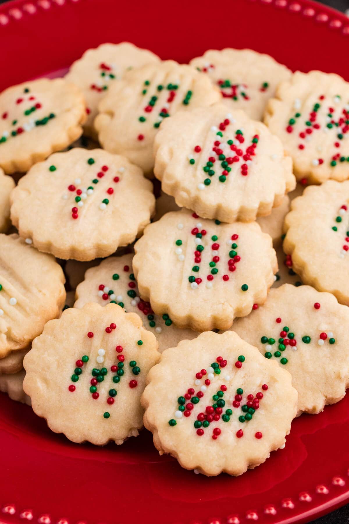 Round holiday cookies with scalloped edges and colorful red, green, and white sprinkles, stacked on a red ceramic plate, perfect for festive Christmas gatherings.