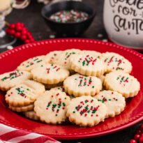 Holiday cookies with scalloped edges topped with red, green, and white sprinkles, arranged on a red plate surrounded by festive decor, including a striped napkin, red berries, and a "Cookies for Santa" jar.