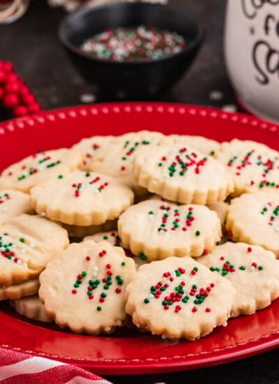 Holiday cookies with scalloped edges topped with red, green, and white sprinkles, arranged on a red plate surrounded by festive decor, including a striped napkin, red berries, and a "Cookies for Santa" jar.