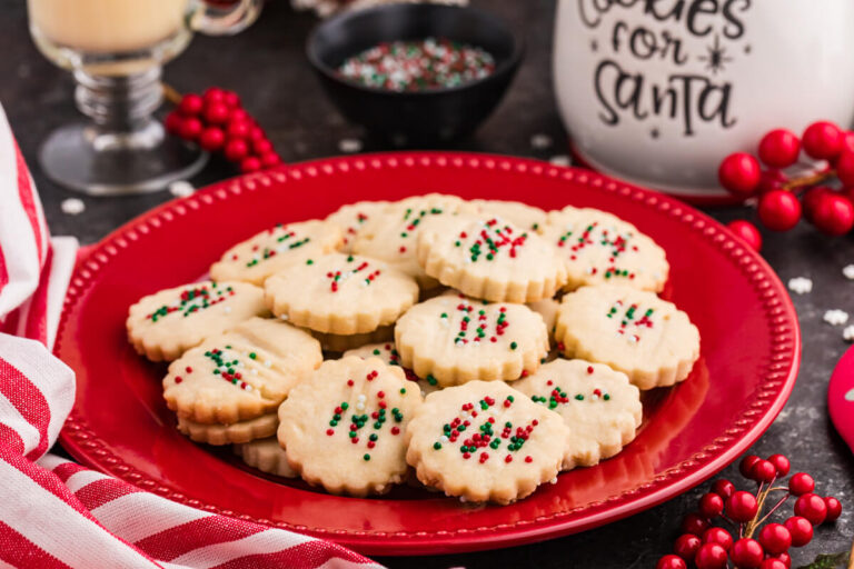 Holiday cookies with scalloped edges topped with red, green, and white sprinkles, arranged on a red plate surrounded by festive decor, including a striped napkin, red berries, and a "Cookies for Santa" jar.