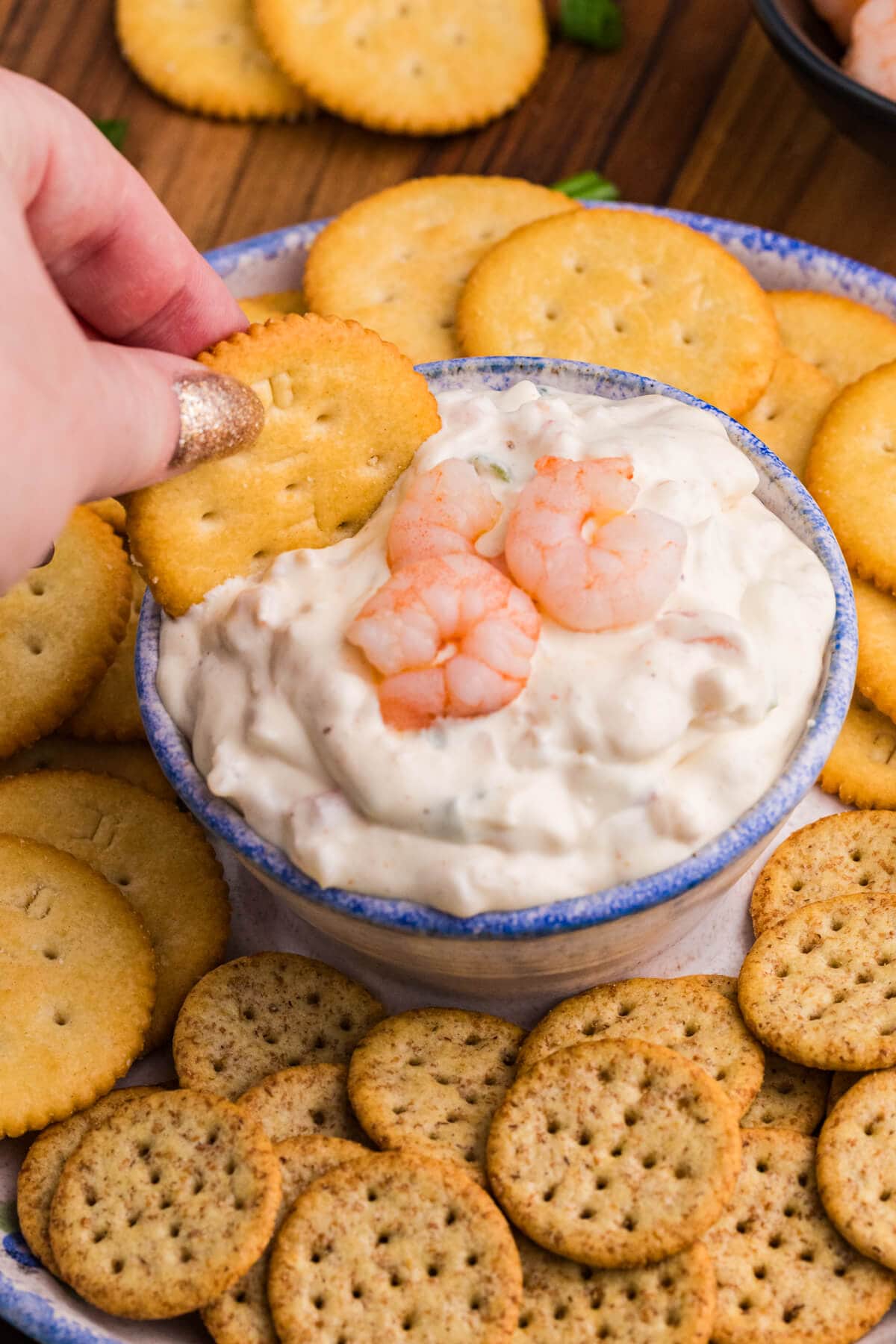 Hand dipping a Ritz cracker into shrimp-topped creamy dip, surrounded by assorted crackers.