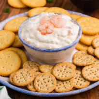 Creamy dip topped with shrimp in a ceramic bowl, served with Ritz and multigrain crackers on a rustic plate.