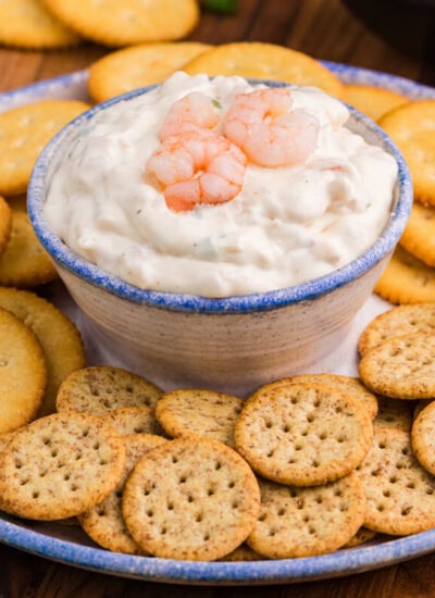 Creamy dip topped with shrimp in a ceramic bowl, served with Ritz and multigrain crackers on a rustic plate.