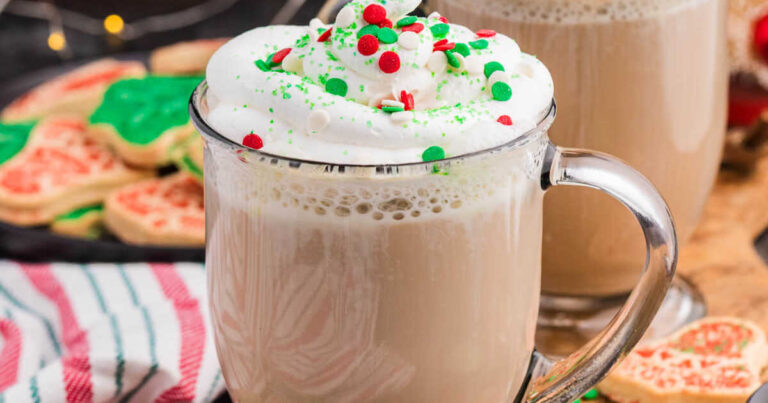 Glass mug filled with a frothy holiday latte, topped with whipped cream, festive red, green, and white sprinkles, and a green sugar-coated Christmas tree cookie resting on top, with more decorated cookies and a striped towel in the background.