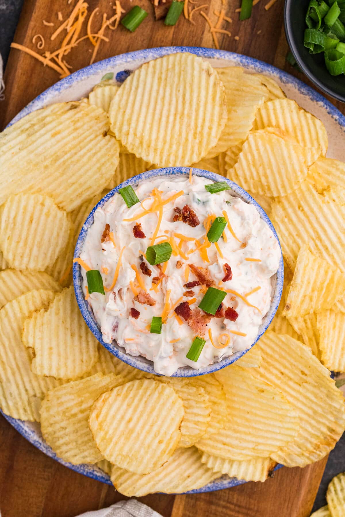 Crack Dip topped with bacon, cheddar, and green onions, served with ridged potato chips on a wooden board.