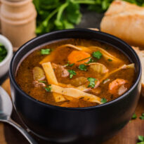Bowl of beef and noodle soup with vegetables, garnished with parsley, served with bread.
