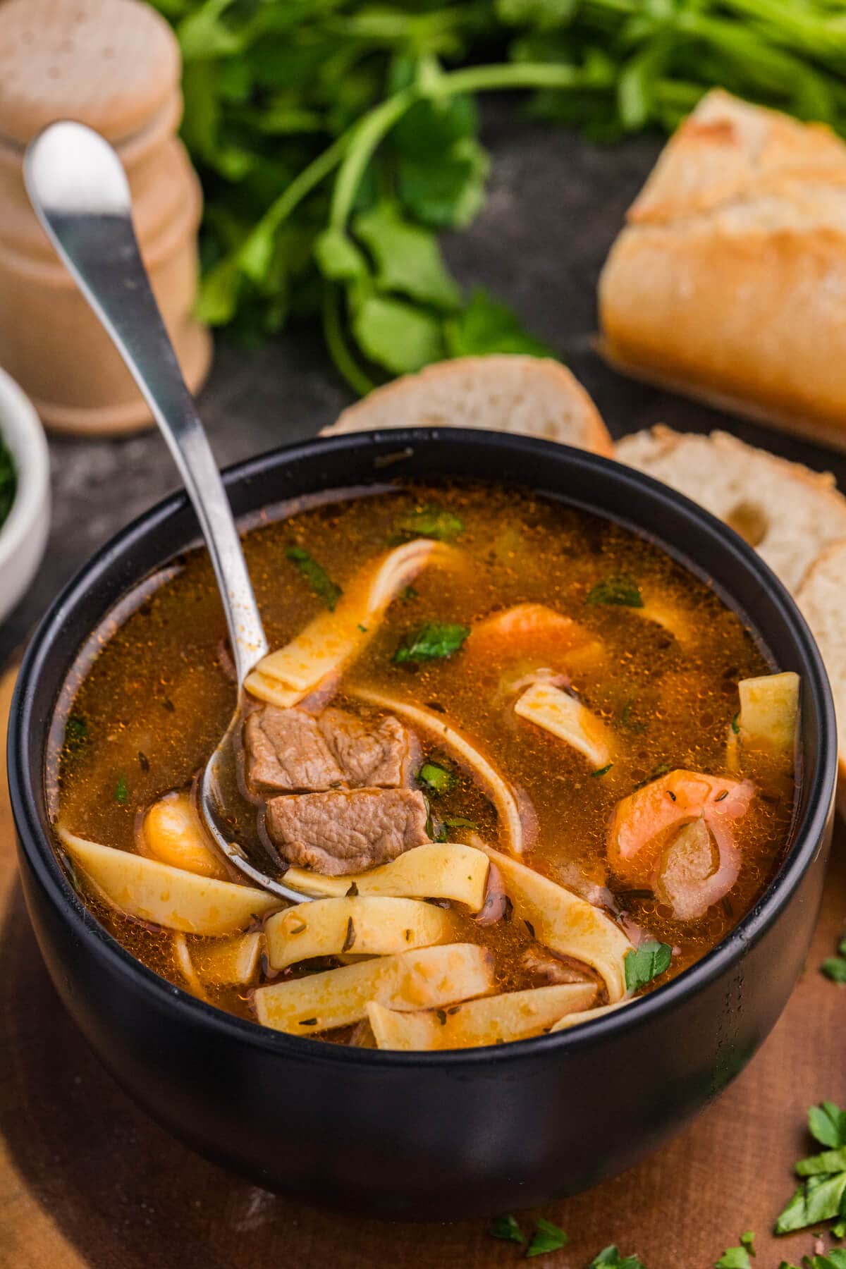 Spoonful of beef noodle soup with carrots and noodles in a black bowl, fresh bread in background.