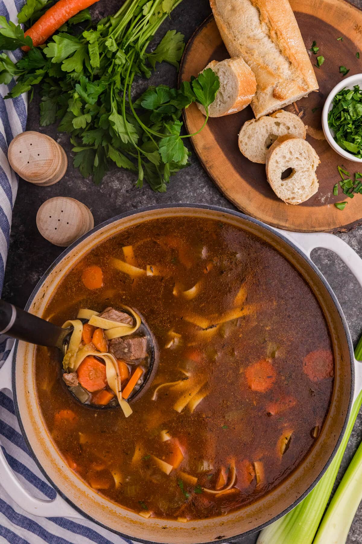 Pot of beef noodle soup with beef chunks, egg noodles, carrots, and celery in broth, surrounded by bread, parsley, and seasoning.