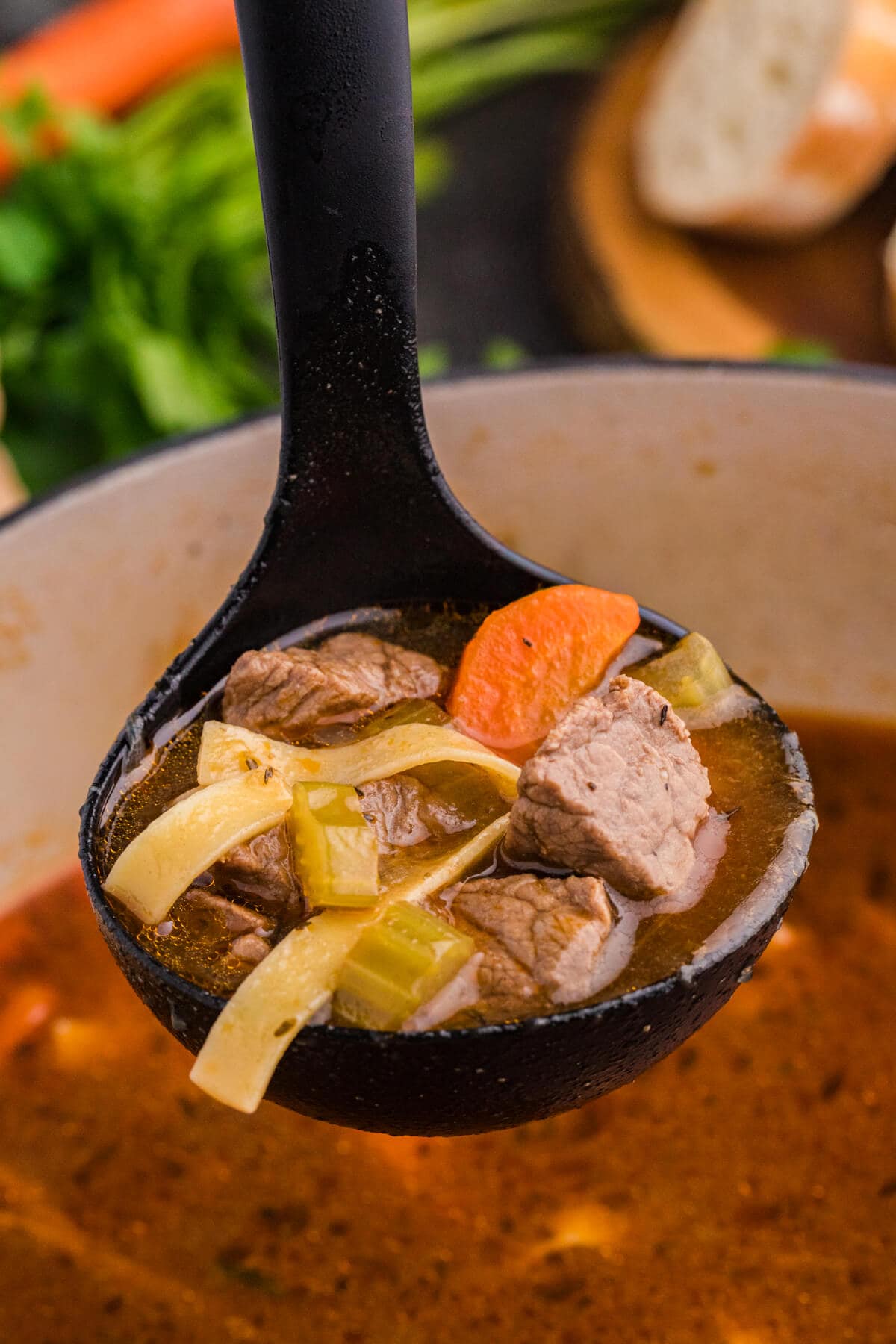 Ladle lifting beef, noodles, carrots, and celery from a hot soup pot.