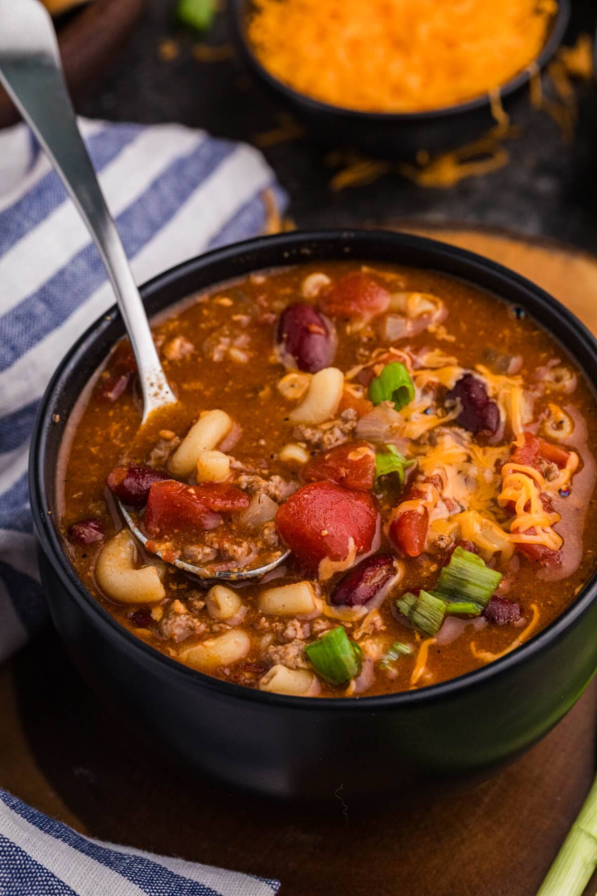 Spoon lifting macaroni, ground beef, kidney beans, tomatoes, and melted cheddar from a black bowl of savory tomato-based soup.