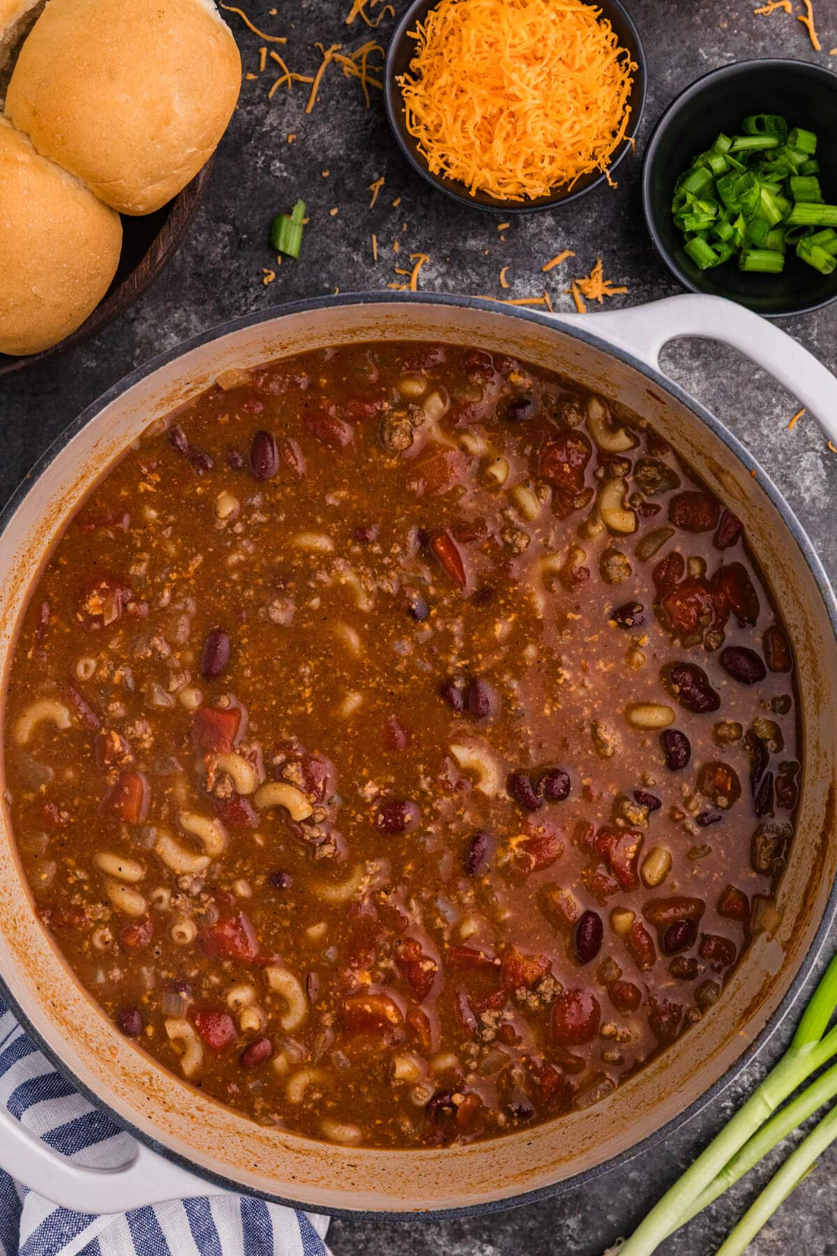 Chili Mac Soup in a white Dutch oven with elbow macaroni, ground beef, kidney beans, and tomatoes, served with shredded cheddar, green onions, and dinner rolls.