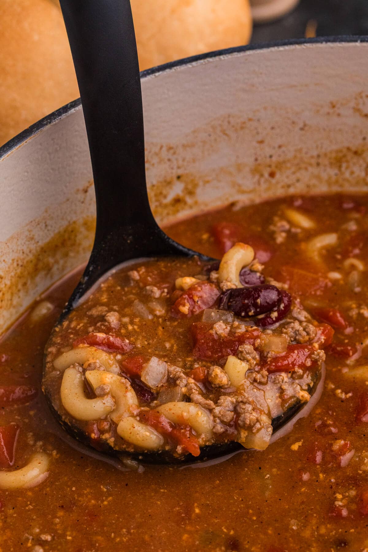 Close-up of a ladle scooping hearty macaroni, ground beef, kidney beans, and tomato broth from a white Dutch oven.