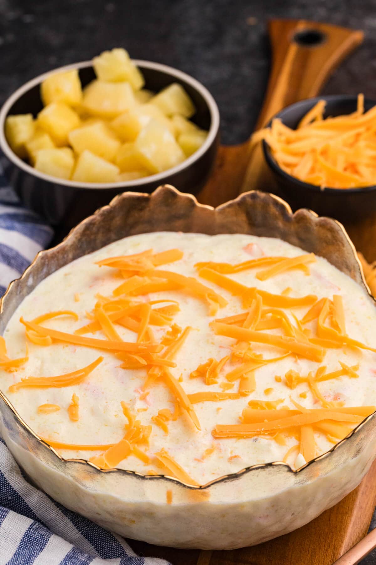 Pineapple salad topped with shredded cheddar cheese, served in a decorative glass bowl, with small bowls of pineapple chunks and grated cheese in the background on a wooden board.
