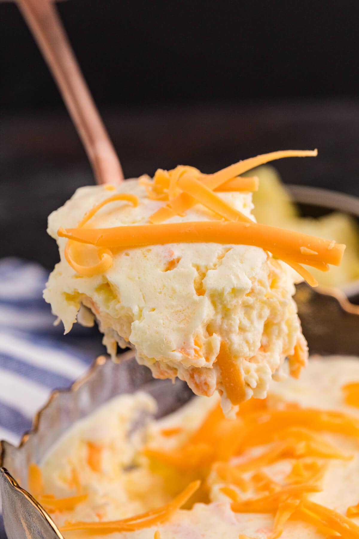 Close-up of a spoonful of creamy fruit salad with visible pineapple chunks and shredded cheddar cheese, lifted above the main serving dish.