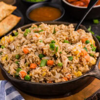 Chicken fried rice in a skillet, garnished with green onions, surrounded by egg rolls and sweet sauces.